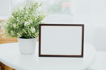 A blank picture frame on a table beside a decorative potted plant in a bright room