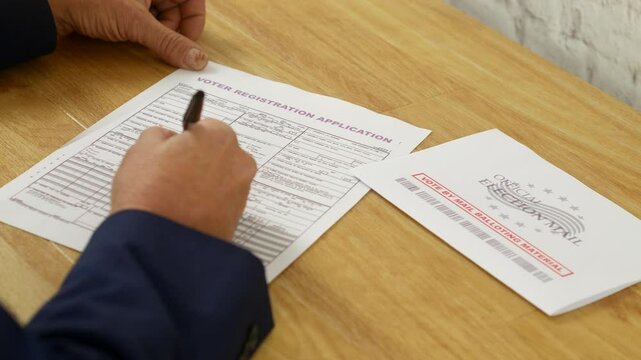 A man filling out a voter registration application with a pen on a wooden table. An "Official Election Mail" envelope rests nearby, emphasizing the process of registering to vote by mail