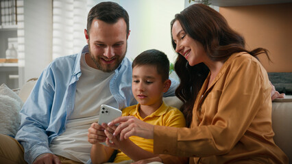 Happy Caucasian family man woman father mother kid boy child son making purchase in online toy store browsing social network make order in Internet on phone smartphone on couch home parenthood concept