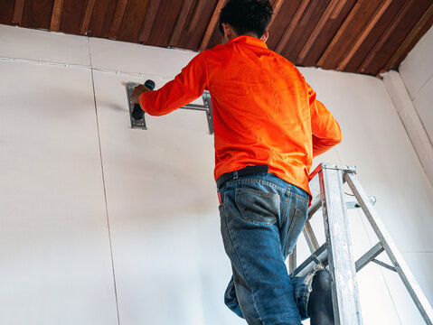 A worker on a ladder uses an electric drill to drill into a white wall to install a Fan Coil Unit: FCU.