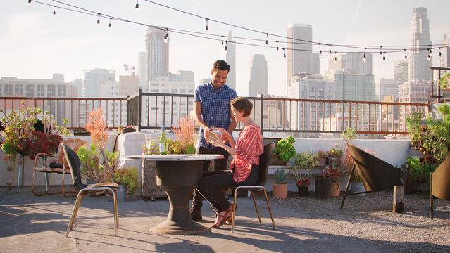 Man Giving Woman Gift As They Celebrate On Rooftop Terrace With City Skyline In Background