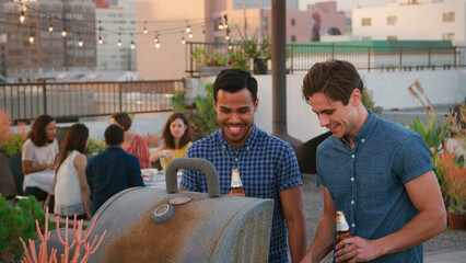 Two Men Cooking Barbecue For Friends Gathered On Rooftop Terrace With City Skyline In Background 