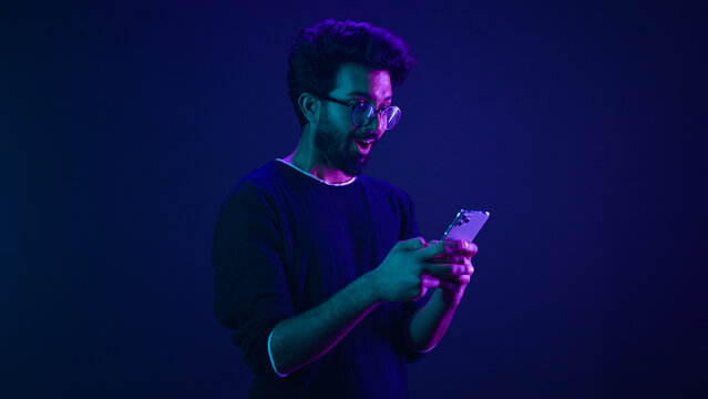Indian man Arabian male model guy win mobile phone success victory smartphone achievement winning game cellphone internet betting online money prize at ultraviolet neon dark blue studio background