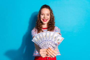 Joyful young woman holding a fan of dollar bills against a vibrant blue background