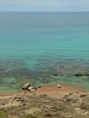 Obraz premium Beach with turquoise waters and rocky shoreline, with beachgoers under an umbrella. Sicily, Italy
