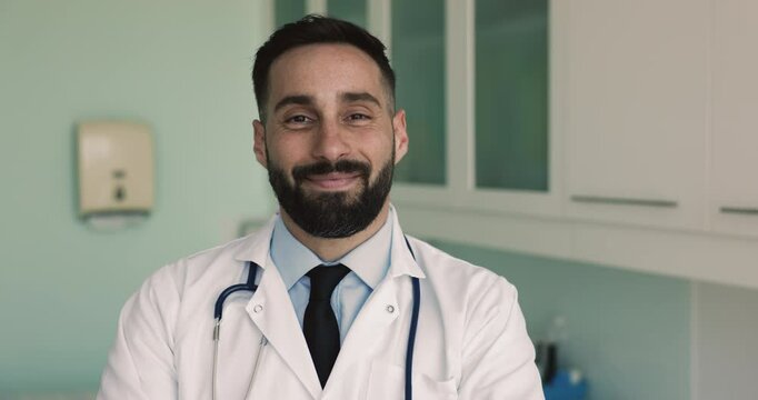 Head shot portrait of handsome smiling Hispanic general practitioner, male internist or cardiologist wear white coat and stethoscope looks at camera posing alone at workplace, Health-care, competence