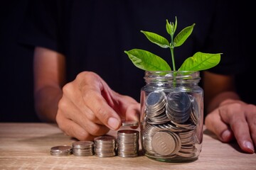Man stacking and counting coins on the wooden table and a plant growing from coins in glass jar. Savings and investment concept.