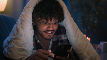 Smiling guy Indian man Arabian male at night evening home on couch under blanket cover with duvet...