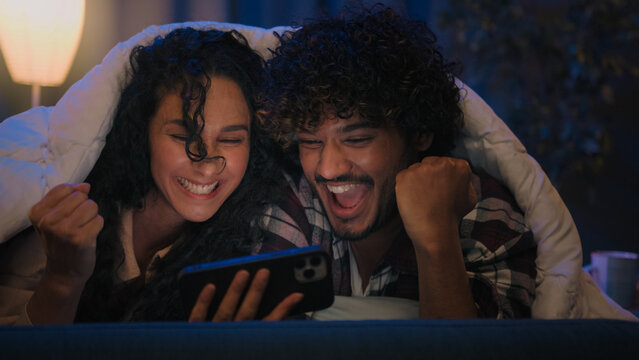 Multiracial couple man and woman at night home win mobile phone bet good news achievement triumph excited with online winning victory smartphone high five at evening couch under blanket gadget addict