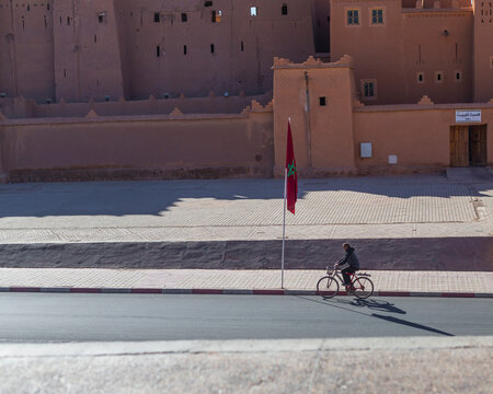 Ouarzazate, Morocco - 18 November 2023: View of traditional adobe buildings with a cyclist and Moroccan flag under sunlight, Ouarzazate, Morocco.
