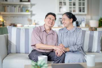 A cheerful elderly couple sitting together on a cozy sofa, holding hands and smiling warmly. They enjoy their peaceful moments together, embodying love, companionship, and happiness in their home.