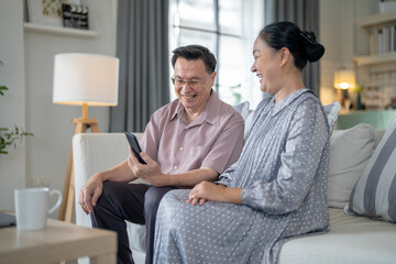 Obraz premium An elderly couple sits together in their cozy living room, smiling and enjoying a moment as they look at something on a smartphone. Their warmth and affection capture a peaceful home atmosphere.