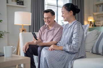 Obraz premium An elderly couple sits together in their cozy living room, smiling and enjoying a moment as they look at something on a smartphone. Their warmth and affection capture a peaceful home atmosphere.