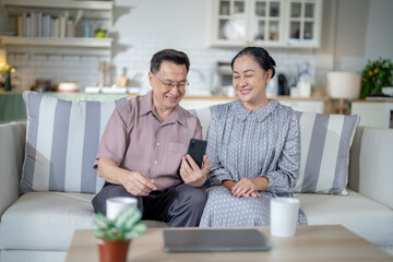 Obraz premium An elderly couple sits together in their cozy living room, smiling and enjoying a moment as they look at something on a smartphone. Their warmth and affection capture a peaceful home atmosphere.