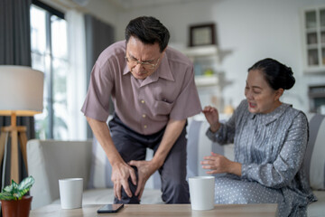 An elderly couple sits in their living room. The man holds his knee, appearing to be in discomfort, while his partner offers support. The image conveys care, concern, and the bond between the couple.