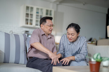 Obraz premium An elderly couple sits in their living room. The man holds his knee, appearing to be in discomfort, while his partner offers support. The image conveys care, concern, and the bond between the couple.