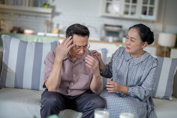 An elderly couple sits on a couch in a cozy living room. The man holds his head in discomfort, while his partner offers support. The image highlights care, concern, and the bond between the couple.