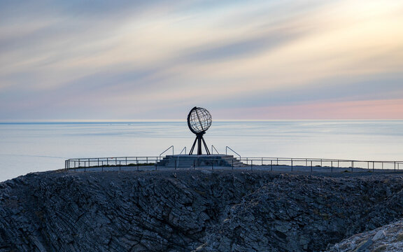 View of the iconic globe sculpture on a rocky cliff overlooking the serene ocean at sunset, Nordkapp, Norway.