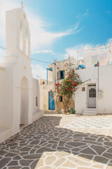 A tranquil Greek courtyard features beautifully whitewashed buildings and a small church with a bell tower, surrounded by vibrant plants under bright blue skies.