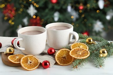 Tasty hot cocoa drinks in cups, fir branches, baubles and dry orange slices on white table against blurred lights