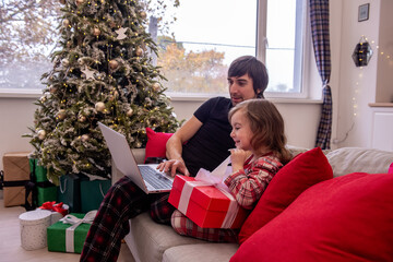 Young father and daughter in plaid pajamas enjoy heartwarming moment on sofa, with laptop, wrapped red gift, surrounded by holiday cheer, decorated Christmas tree. Shopping online, sales