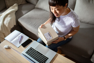 Smiling Asian woman opening package at home with laptop