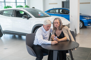 A mature couple is sitting in a car dealership and looking at a smartphone. 