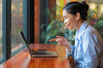 excited asian woman sitting in cafe talking on laptop have video chat, online meeting, with pointing finger wearing striped blue shirt