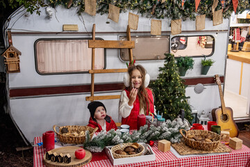 Two children dressed warmly sit at a festive outdoor table with snacks, surrounded by holiday decor, including Christmas lights and a tree, in front of a cozy caravan.