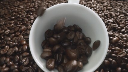 Close up of coffee bean falling in to coffee cup surrounded by pile of beans. Dropping aromatic blending bean in to white cup. Top view of coffee drop in to glass with black background. Comestible.