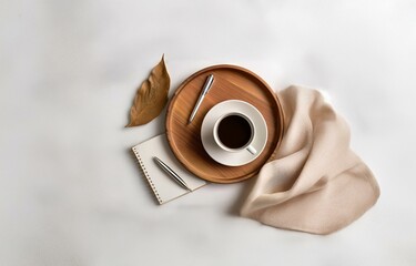 A white surface with a wooden tray holding a cup of coffee, a pen, and a notepad. A single leaf is off to the side of the tray, and a piece of fabric drapes to the right.