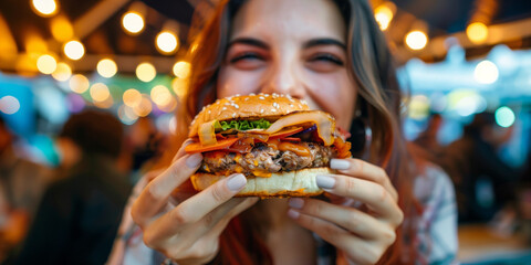 An attractive young woman, smiling cheerfully, holds a tasty burger in two hands. Outdoor street food festival, eat market in the street.