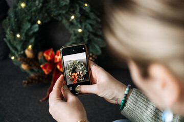 Woman photographing black cat with Christmas wreath and lights for social media content. Festive holiday decorations in cozy home setting.