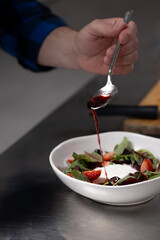 Hand holding spoon and drizzling rich sauce over fresh salad of strawberries and greens on kitchen stainless steel countertop