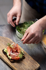 Person carefully arranging slices of fresh strawberries and ham on top of dark bread base spread with avocado