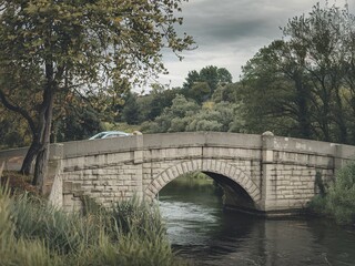 Fototapeta premium A stone bridge over a river with a car on the bridge, surrounded by trees and greenery