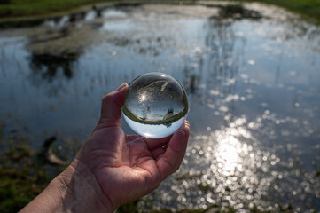 A small lake in green nature is reflected in a ball that a human hand is holding.