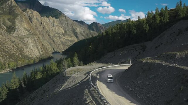 White automobile microvan riding on grail dust road of alpine valley drone shot. Auto travel trip on path surrounded natural sunlight highland cliff rock geology formation