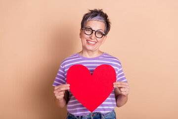 Photo of adorable woman wear striped stylish clothes hand hold red paper card isolated on beige color background
