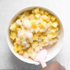 Overhead view of diced pineapple and sugar being mixed together in a white bowl, process of making pineapple cheong or pineapple syrup