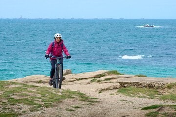 active senior woman cycling with her electric mountain bike at the rocky red granite cost next to Ploumanac'h,  C&ocirc;tes d'Armor,Brittany, France