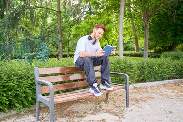 Fototapeta premium A man is sitting on a park bench reading a book. He is wearing headphones and he is enjoying his time. The park is filled with trees and bushes, creating a peaceful and relaxing atmosphere