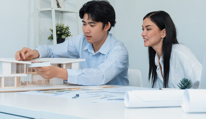 Obraz premium Professional male asian architect using ruler to measure house model length while young beautiful caucasian colleague using laptop to analyzed data on meeting table with house model. Immaculate.