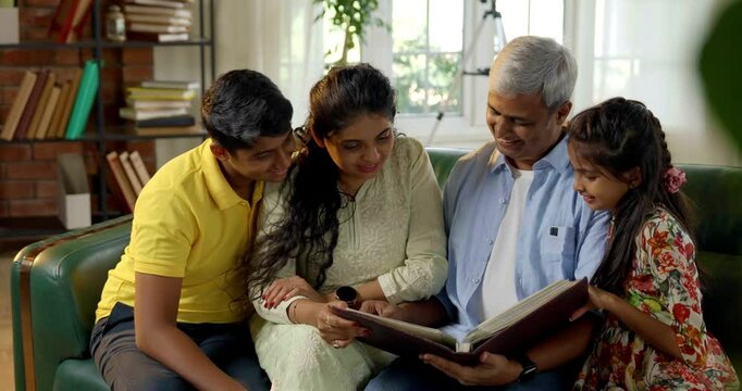 A closeup of a happy Indian Asian family consisting of parents in their mid-forties, a teenage son, and a small daughter joyfully reminiscing together while flipping through a cherished photo album