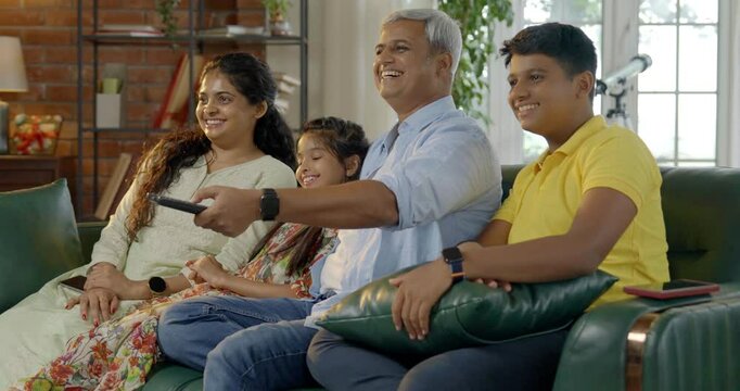 Indian Asian family of four, parents in their forties and two kids, sitting closely on a sofa, laughing out loud while watching television together, enjoying a fun-filled family evening at home