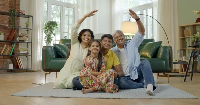 In this close up photo, an Indian Asian family of four,parents in their mid forties, a teenage son, and a small daughter,sit on a cozy carpet, smiling joyfully, creating a roof sign with their hand