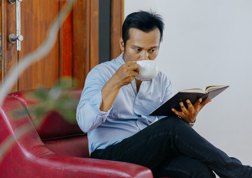 Asian business man drinking a hot coffee and reading notebook while sitting and relaxing at the red leather sofa over comfortable sitting room.