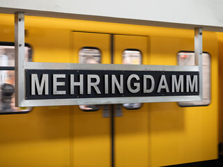 Mehringdamm sign at the underground train station in Berlin Kreuzberg, Germany. Location name of the public transportation. A yellow train is departing in the background.