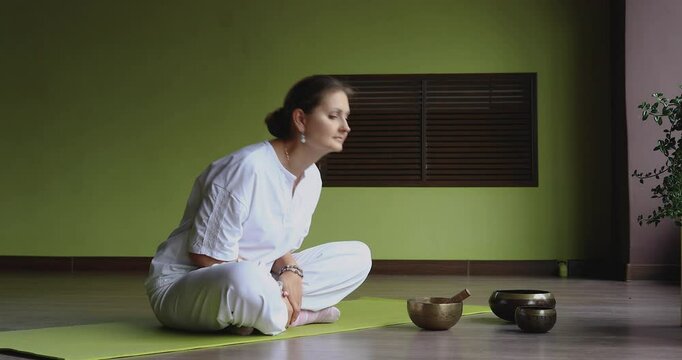 A woman practices Kundalini yoga, does spinal bends while sitting in the lotus position, trains in white comfortable clothes in a room, full-length view