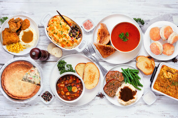Cozy comfort food table scene. Top dwon view over a white wood background. Meatloaf, stew, pot pie, mac and cheese, grilled cheese, soup, fried chicken, shepherds pie and warm sides.